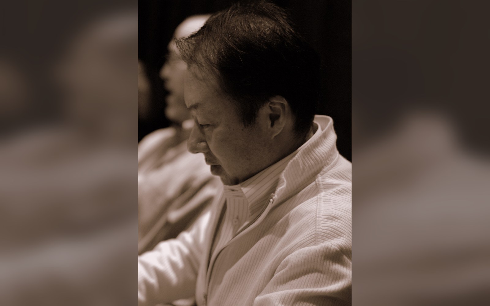 Man in profile wearing a ribbed jacket, seated and looking downward in sepia tone